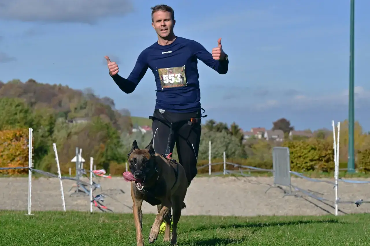 Mężczyzna z psem rasy malinois biegnie w zawodach psich sportów. Oboje zadowoleni, pokazują kciuki w górę.